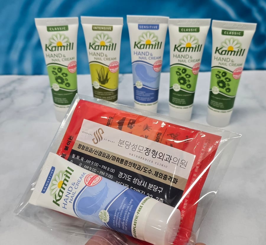카밀 핸드크림 30ml 다섯 개를 뒤에 세워 두고, 앞쪽에 센시티브 핸드크림과 빨간 붙이는 핫팩을 투명 OPP 봉투에 함께 넣은 세트를 들고 있는 모습 — 핫팩 위에는 정형외과 진료 과목·진료 시간·주소를 넣은 홍보 스티커가 붙어 있음